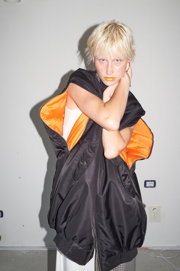 Model wearing the MARIOS black Uovo bomber jacket with arms through the unzipped sleeve openings, revealing the contrasting orange lining.