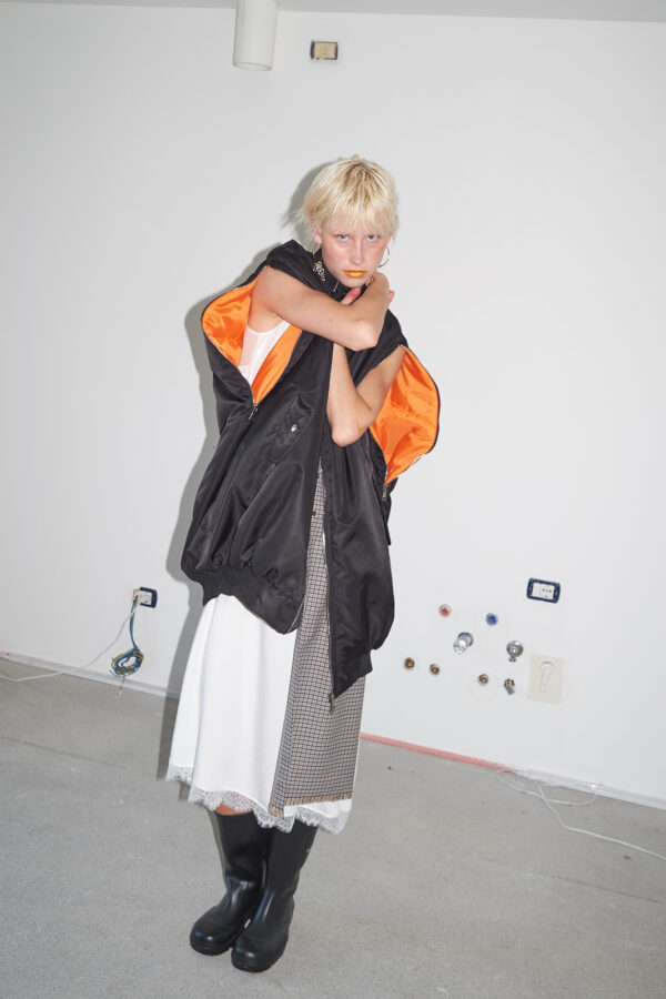 Model wearing the MARIOS black Uovo bomber jacket with arms through the unzipped sleeve openings, revealing the contrasting orange lining.