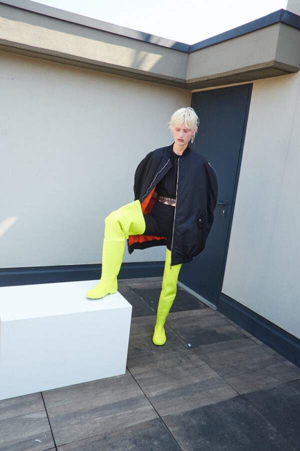 Model wearing the MARIOS black Uovo bomber jacket open to reveal its orange lining, styled with black clothing and neon yellow thigh-high boots on a terrace.