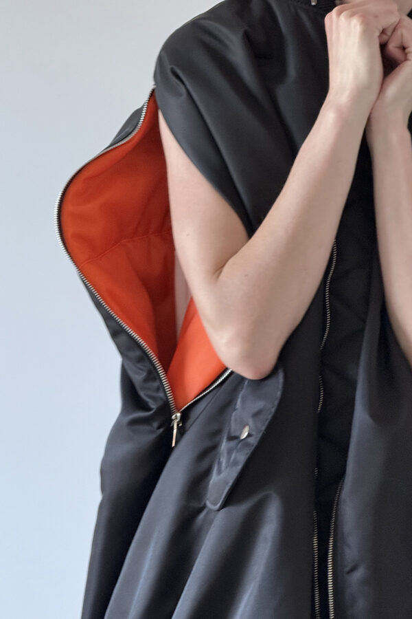 Close-up of the MARIOS black Uovo bomber jacket worn with an arm through the unzipped sleeve opening, revealing the contrasting orange lining and snap-fastening flap pocket.
