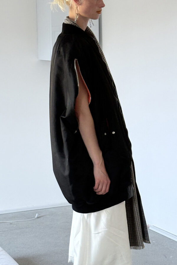 Side view of a model wearing the MARIOS black Uovo bomber jacket, showing its cocoon silhouette and an arm through the adjustable zipped sleeve opening.